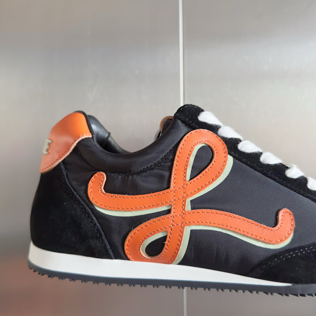 Unisex black sneakers with orange cross emblem, white laces, and rounded toe