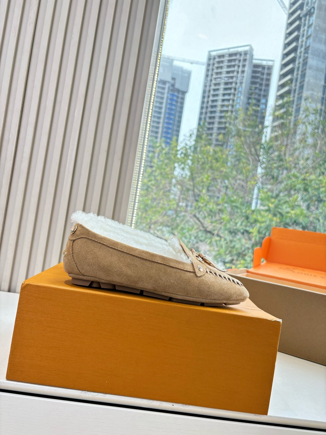 Unisex taupe suede loafer with white shearling lining resting on orange box