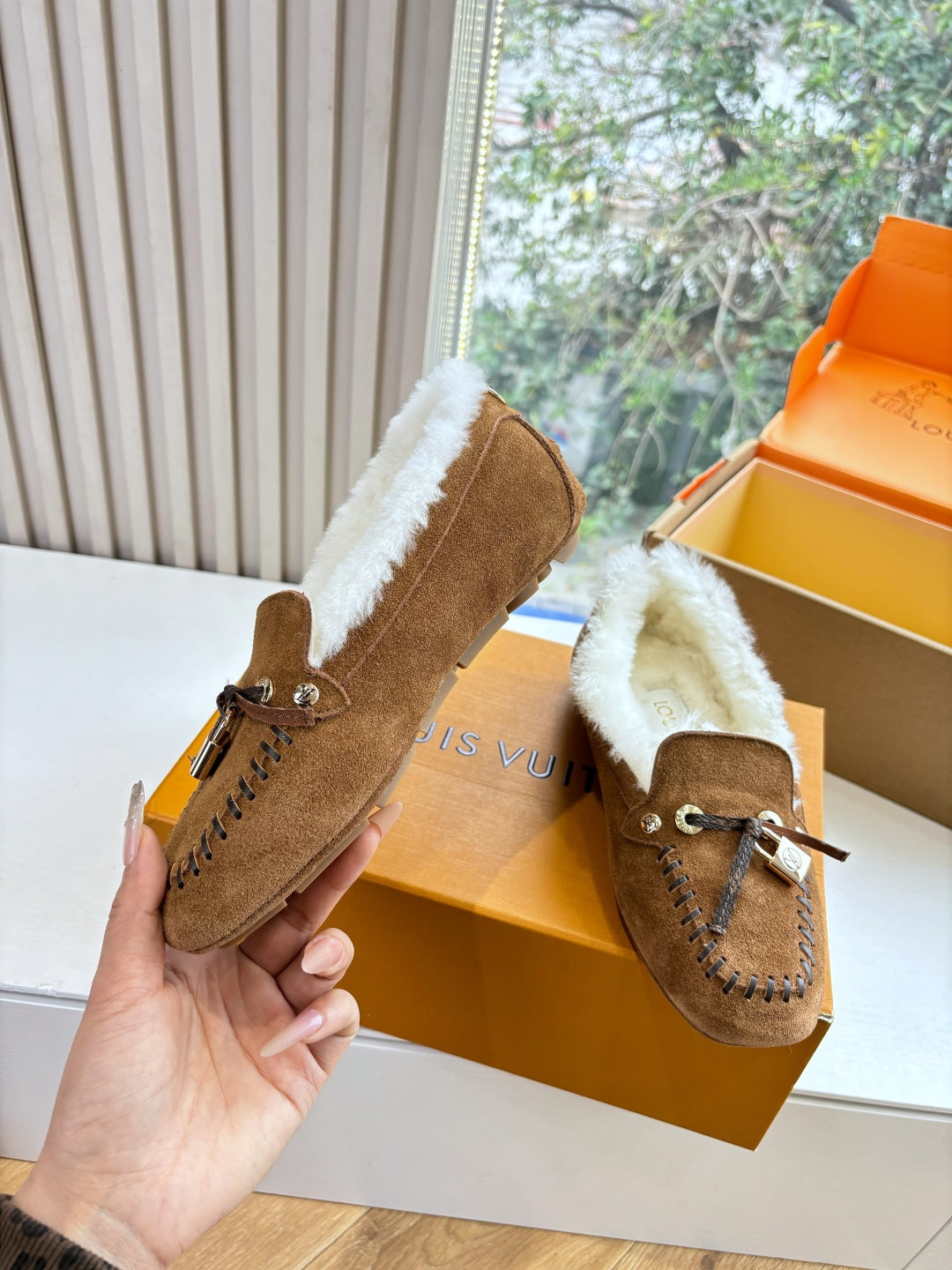 Brown suede shearling loafer in hand, with lace tie, stitched trim, and metal charm