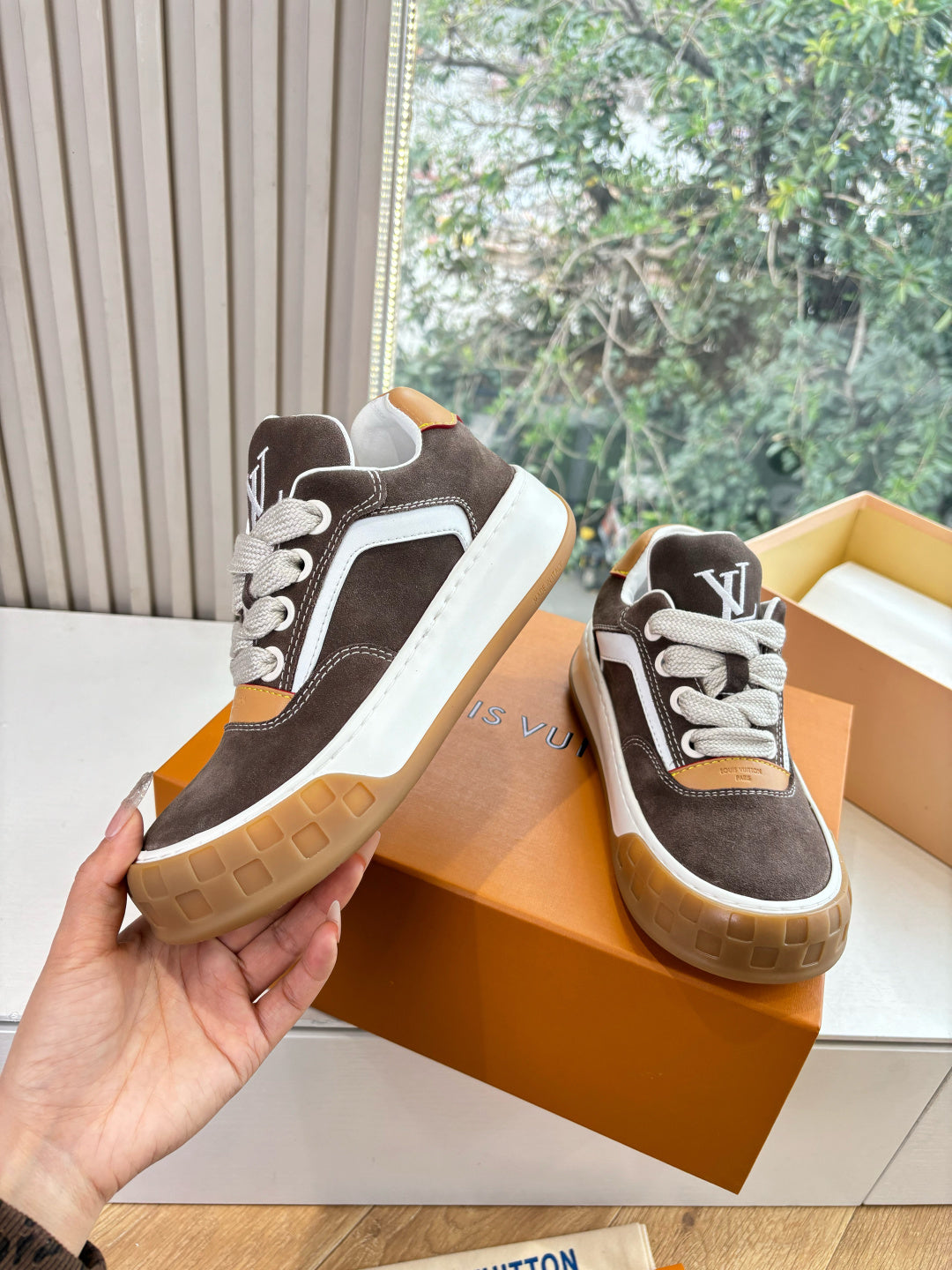 Brown and white sneakers held by hand, LV-style logo patches, tan lace and gum sole.