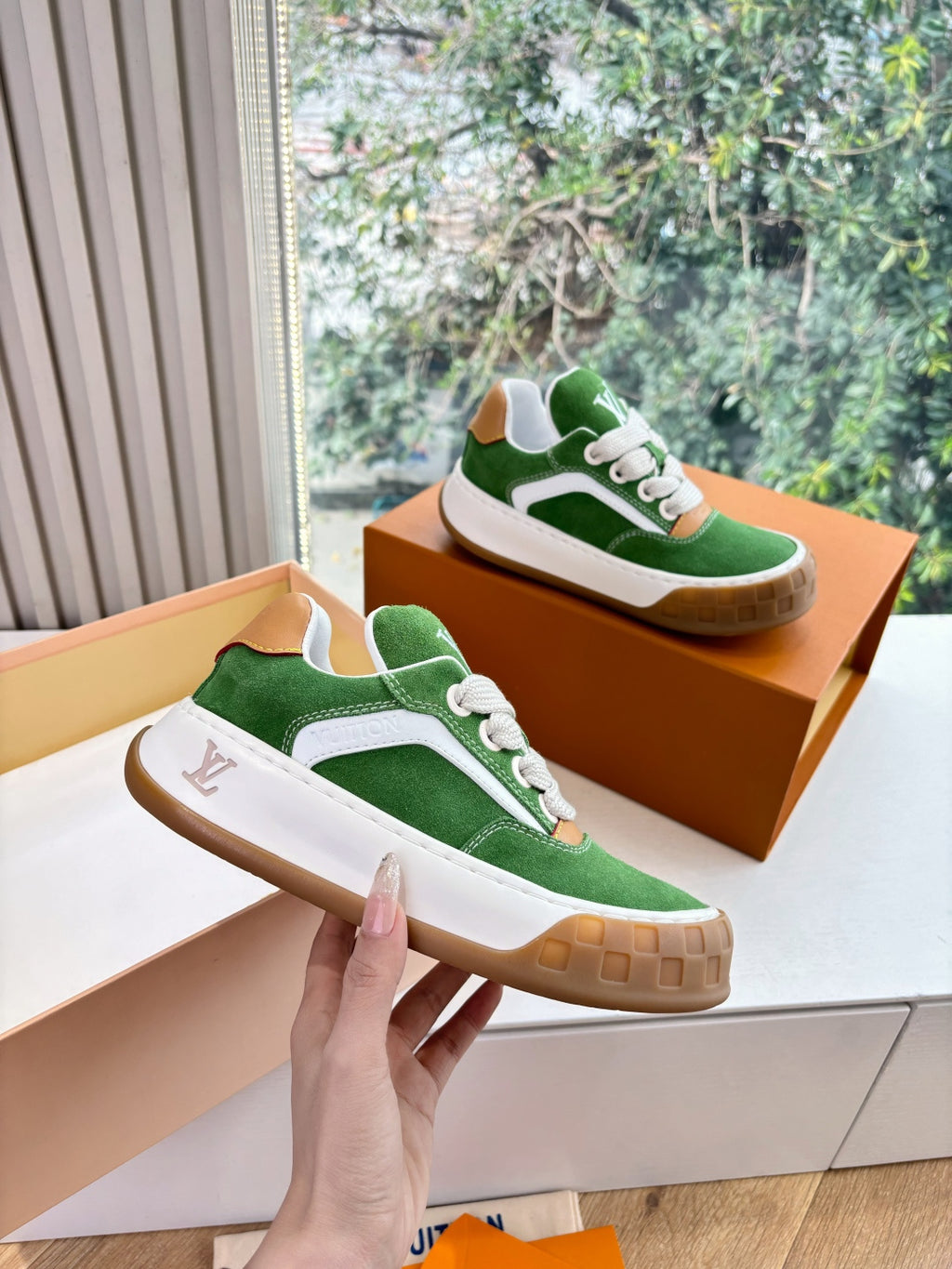 Green suede sneaker pair with white laces, tan sole, held up above orange box