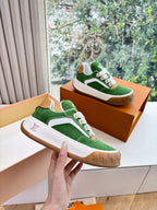 Green suede sneaker pair with white laces, tan sole, held up above orange box
