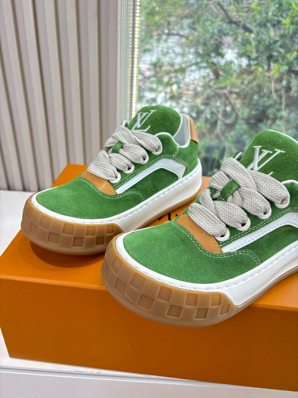 Two green suede sneakers with white laces and tan rubber soles on orange box