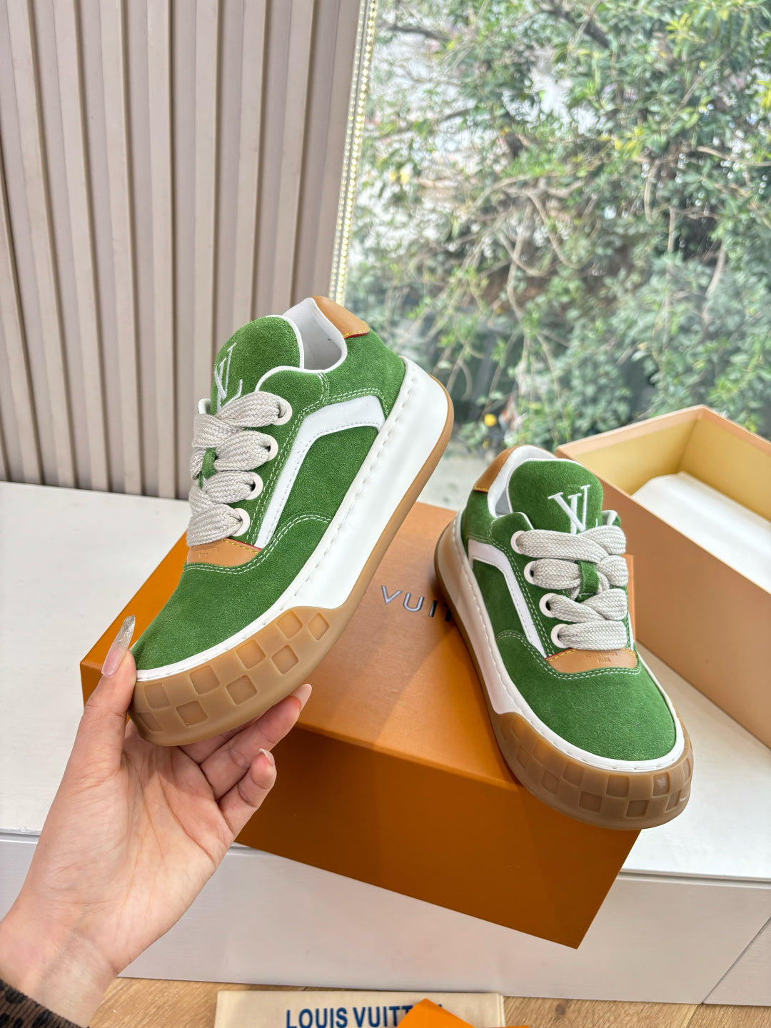 Green suede sneaker held in hand, white laces, tan rubber sole, beside boxed pair