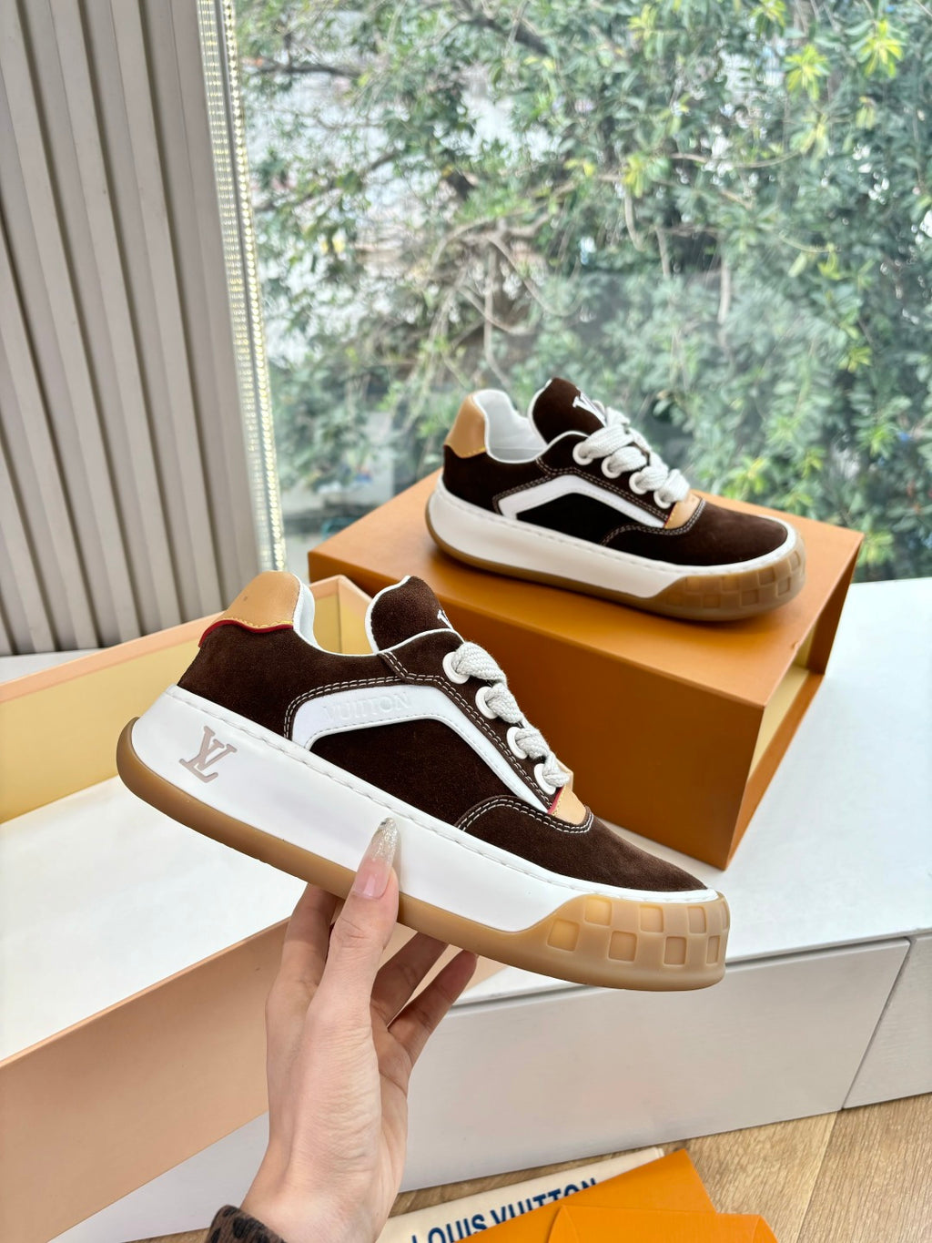 Brown suede sneakers with white sole and LV logo, held in hand above the box.