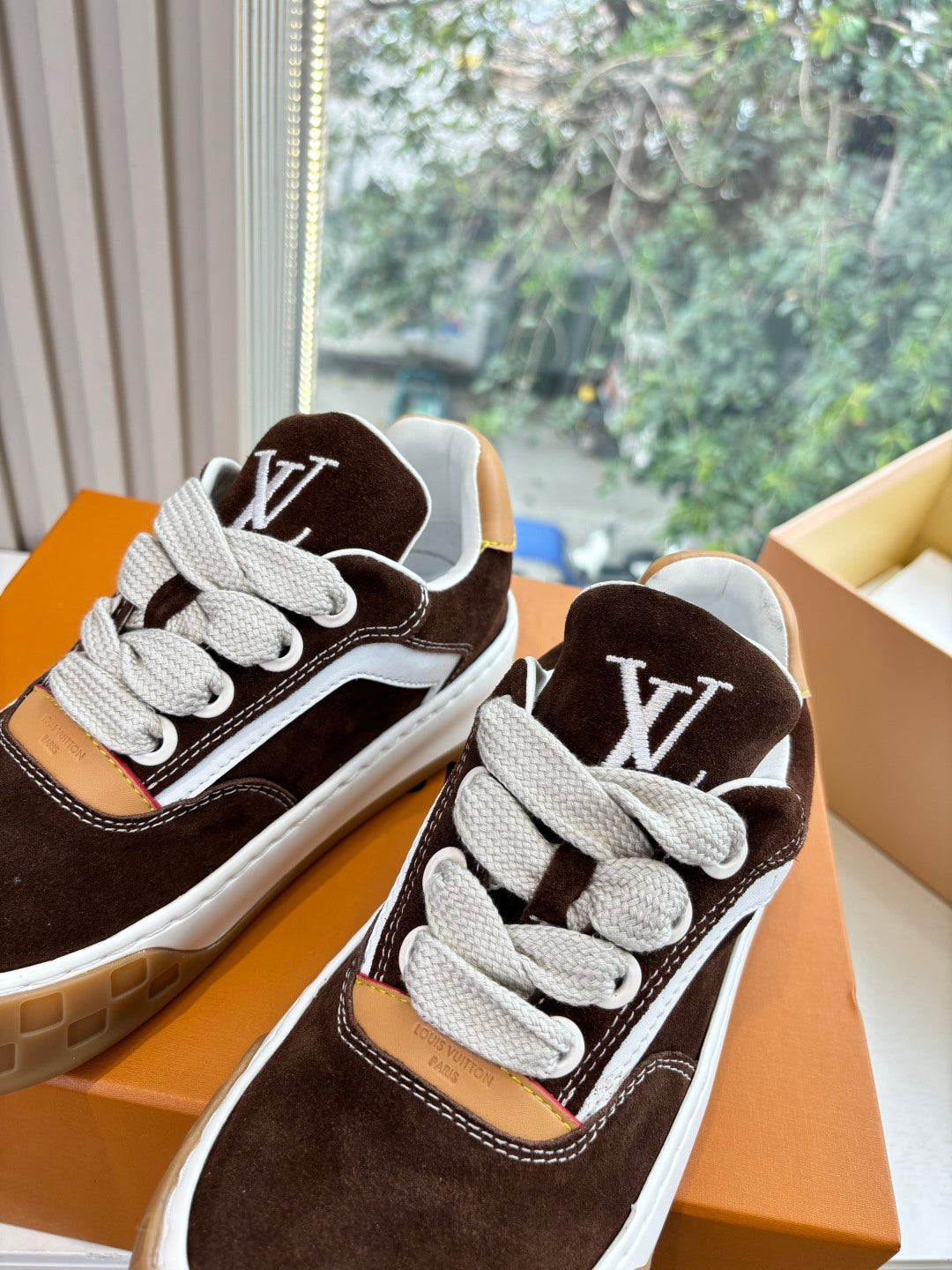 Brown suede sneakers close-up with white laces and LV side emblems, in orange box.