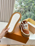 Bottom view of brown suede sneakers with white rubber outsole, in orange box.