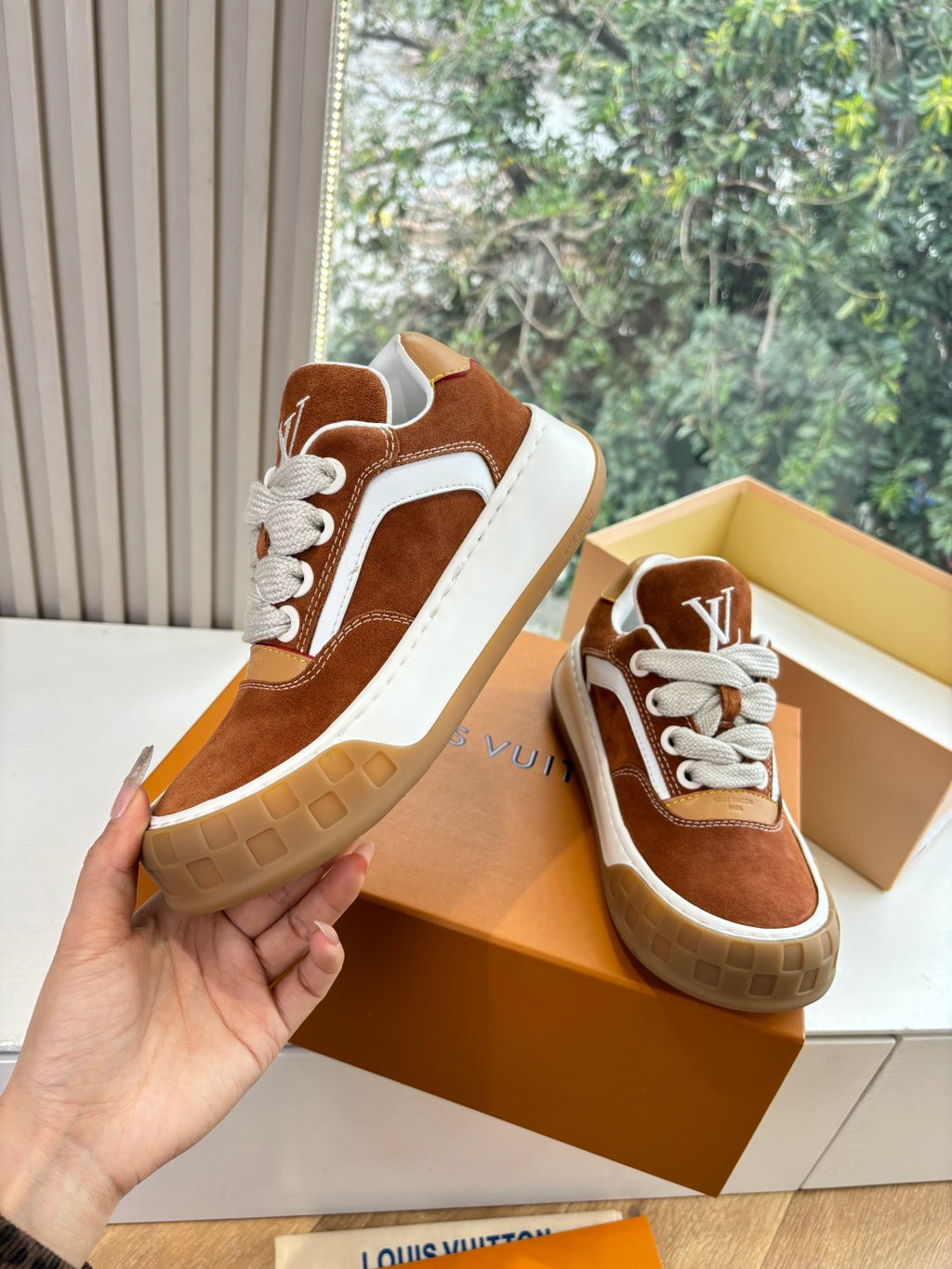 Brown suede low-top sneakers with white laces, held up in orange shoe box.
