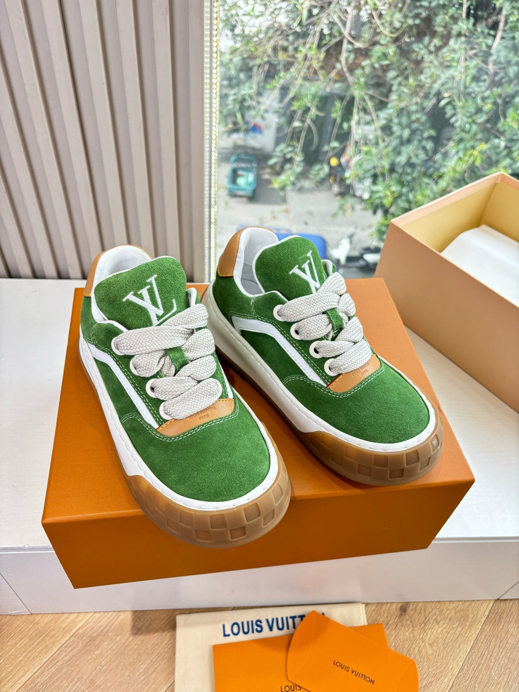 Two green suede sneakers with white laces on orange box insert, tan rubber soles