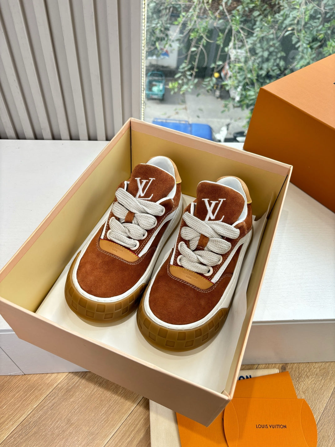 Brown suede sneakers with white laces and LV logo, shown in box.