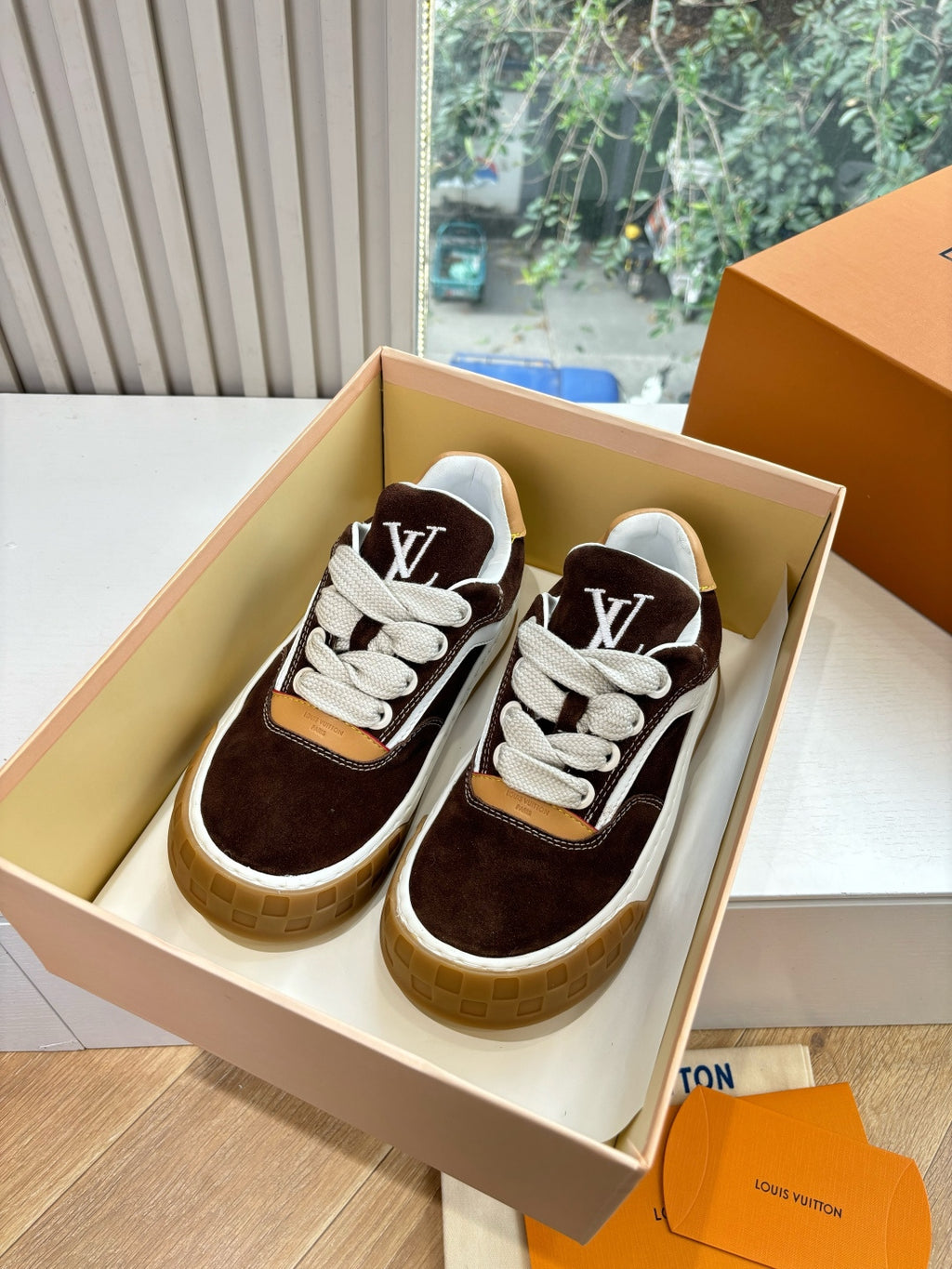 Brown suede sneakers with white laces and LV side emblem, shown in an open box.