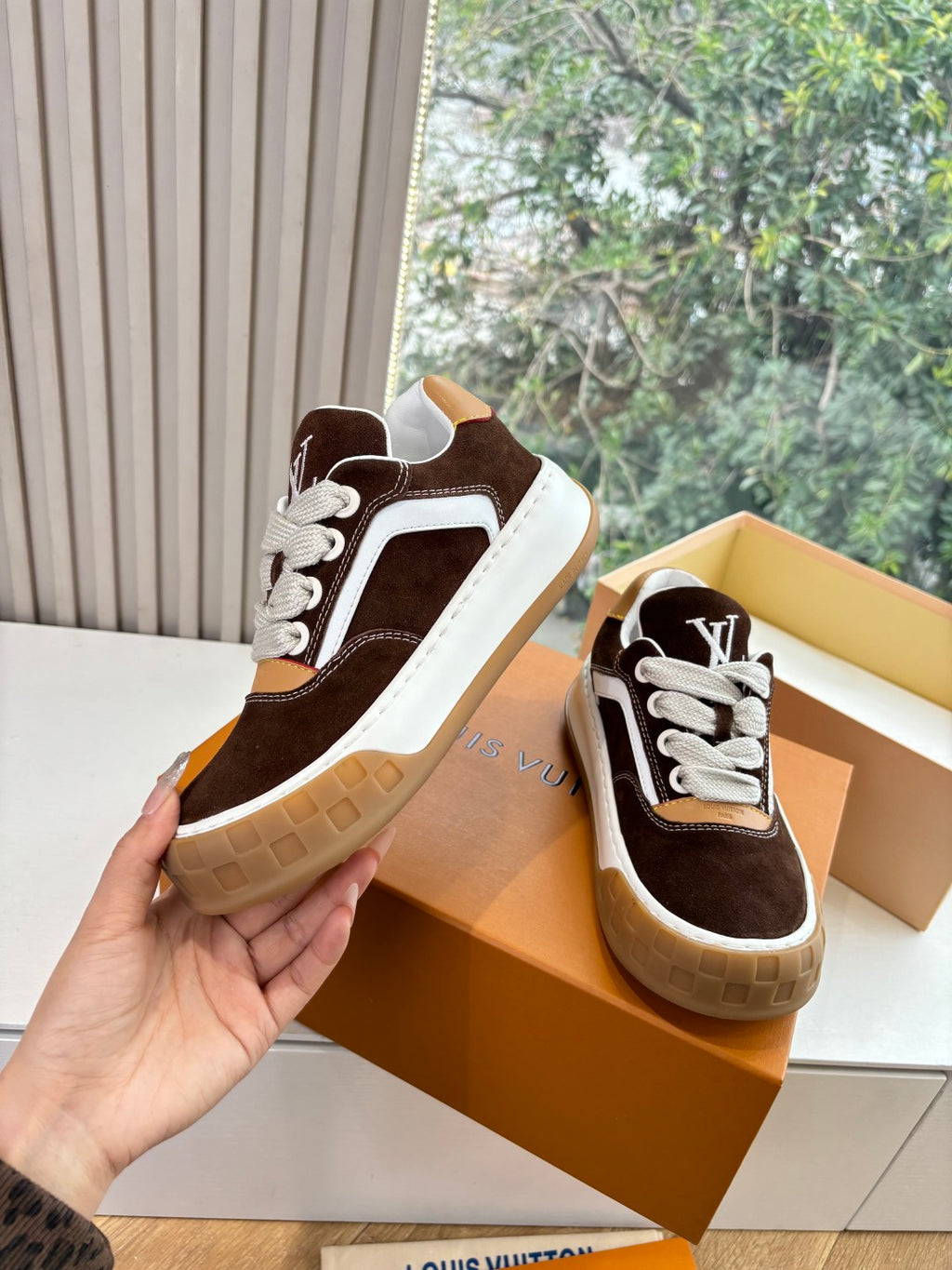 Brown suede sneaker with white trim and LV logo on side, held above orange box.