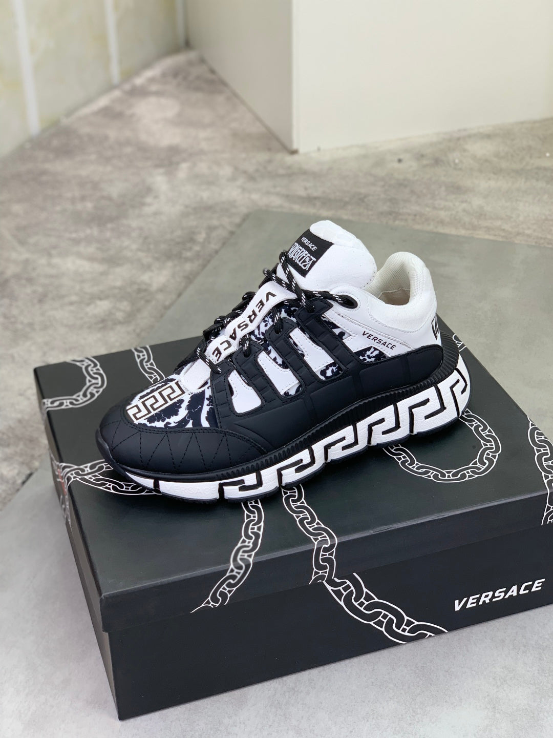 Black sneaker with white laces, white Greca-style trim, and logo patch on side