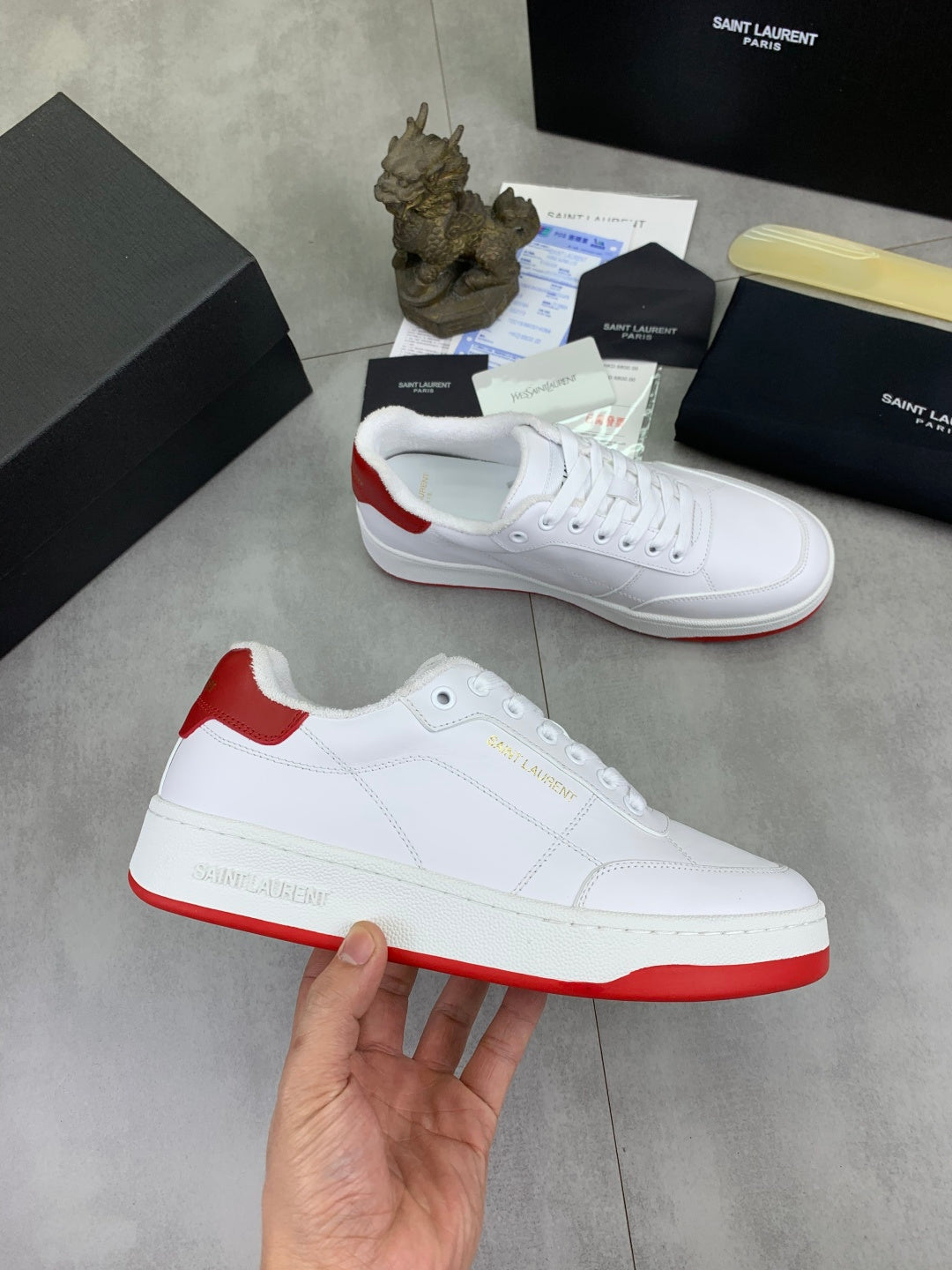 White low-top sneaker in hand, red heel and outsole edge, white lace-up.