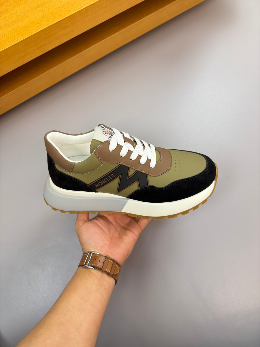 Sneaker held in hand; olive, black, and cream colorblocking with white laces.