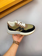 Sneaker held in hand; olive, black, and cream colorblocking with white laces.