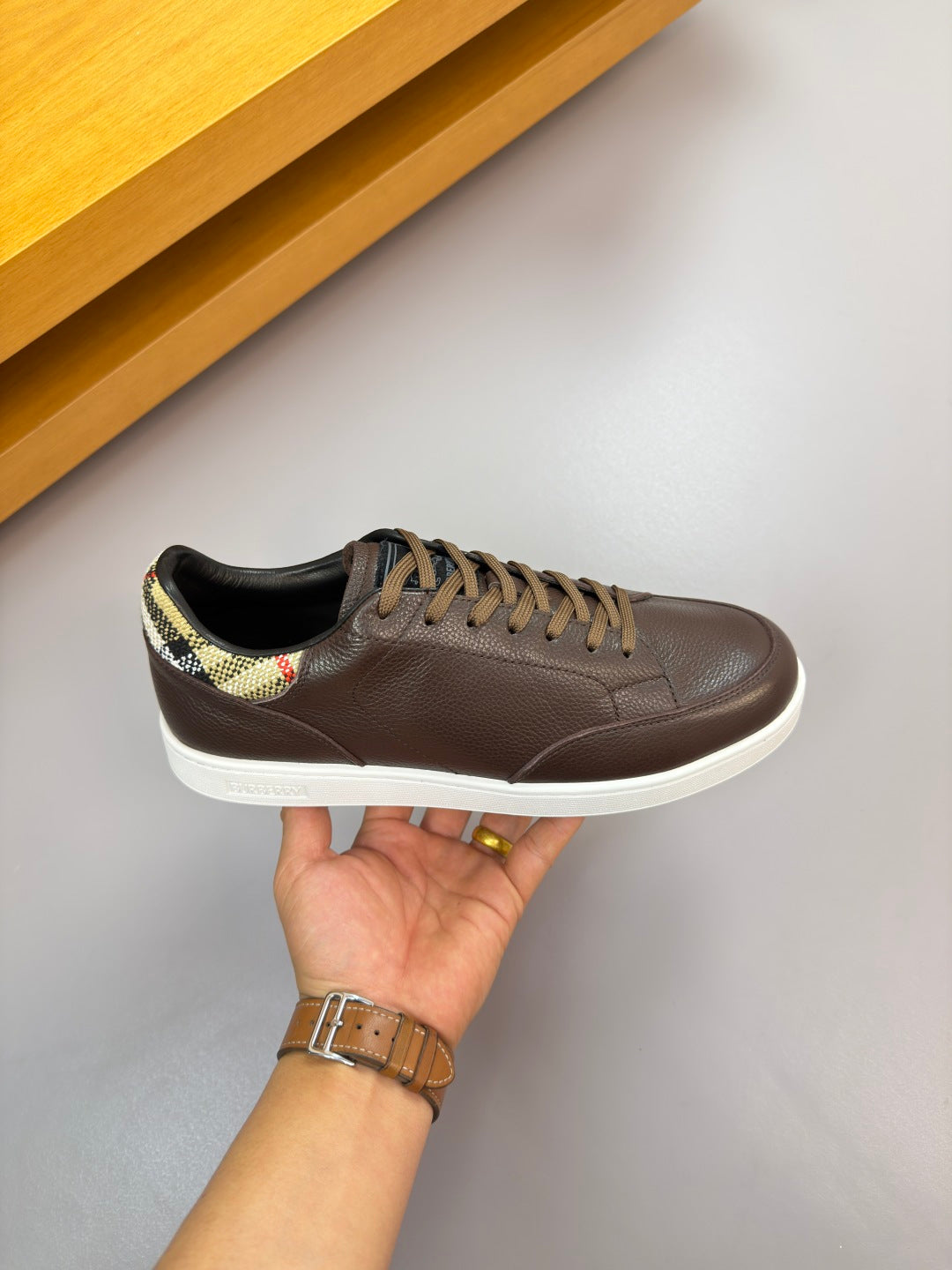 Single brown lace-up sneaker held by hand, with plaid heel accent and white sole