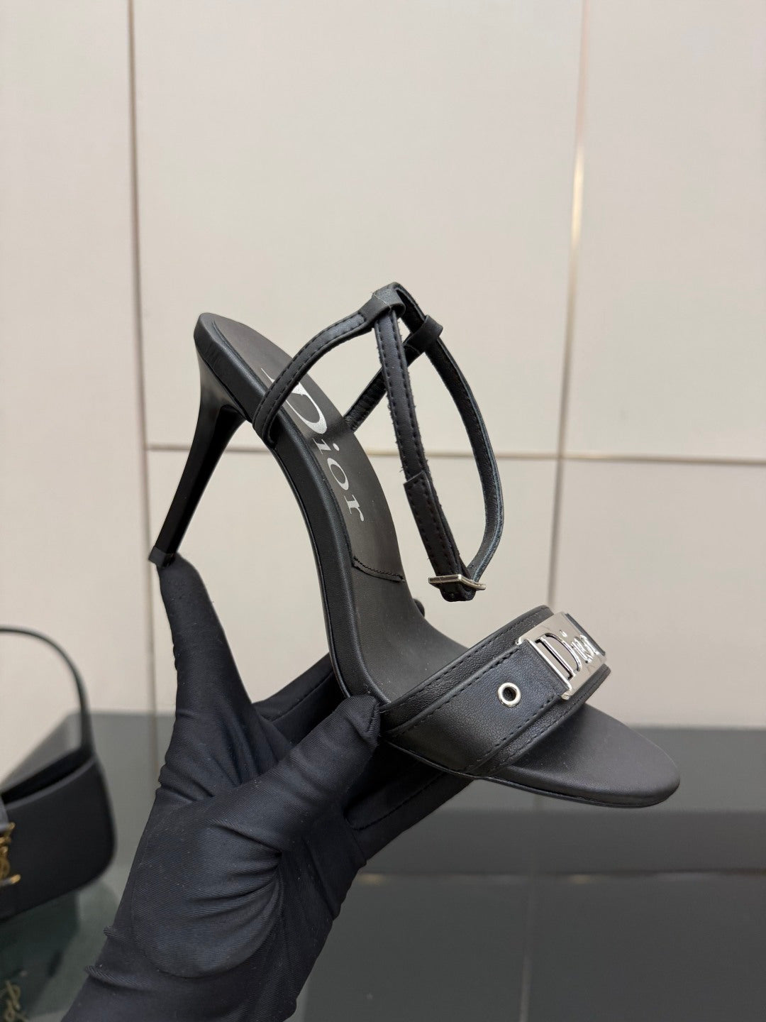 Black leather heeled sandal held in hand; thin straps and Dior label.