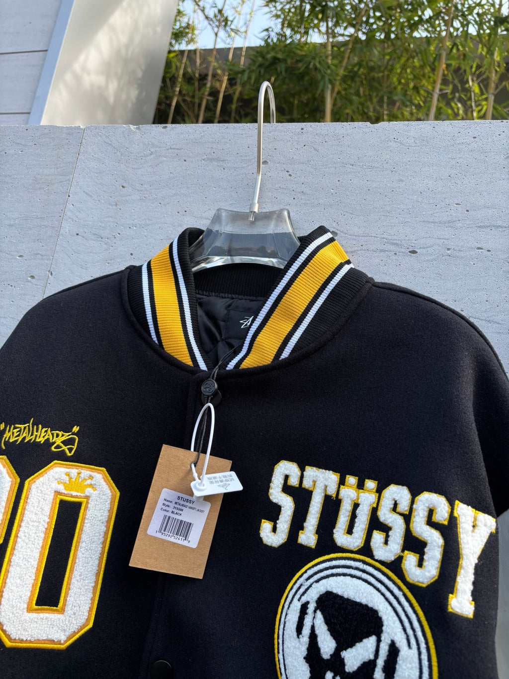 Varsity jacket close-up with Stussy logo, varsity patch emblems, and hanging price tags