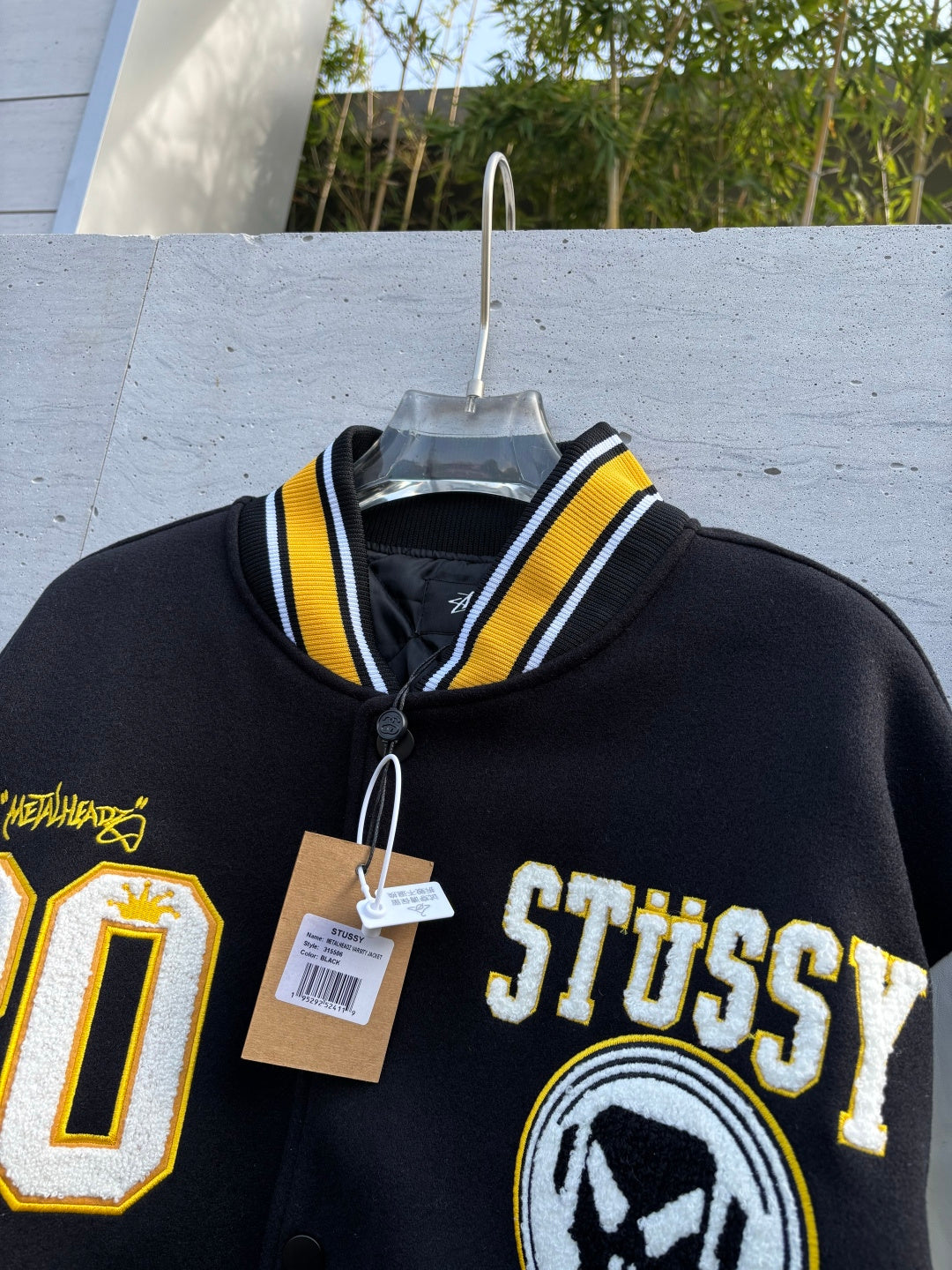 Varsity jacket close-up with Stussy logo, varsity patch emblems, and hanging price tags