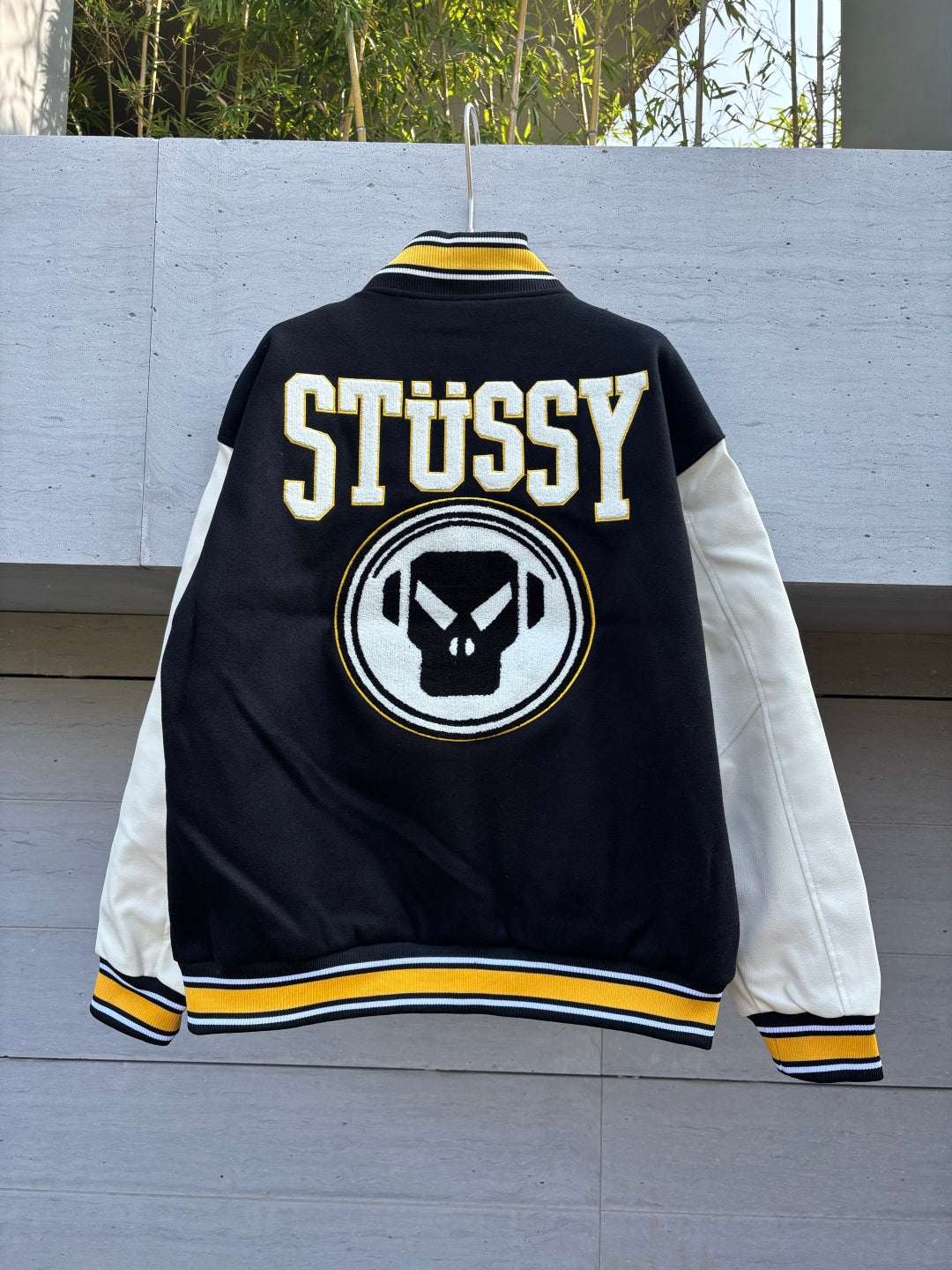 Back varsity jacket with large STUSSY text, skull emblem, and yellow-and-white trim