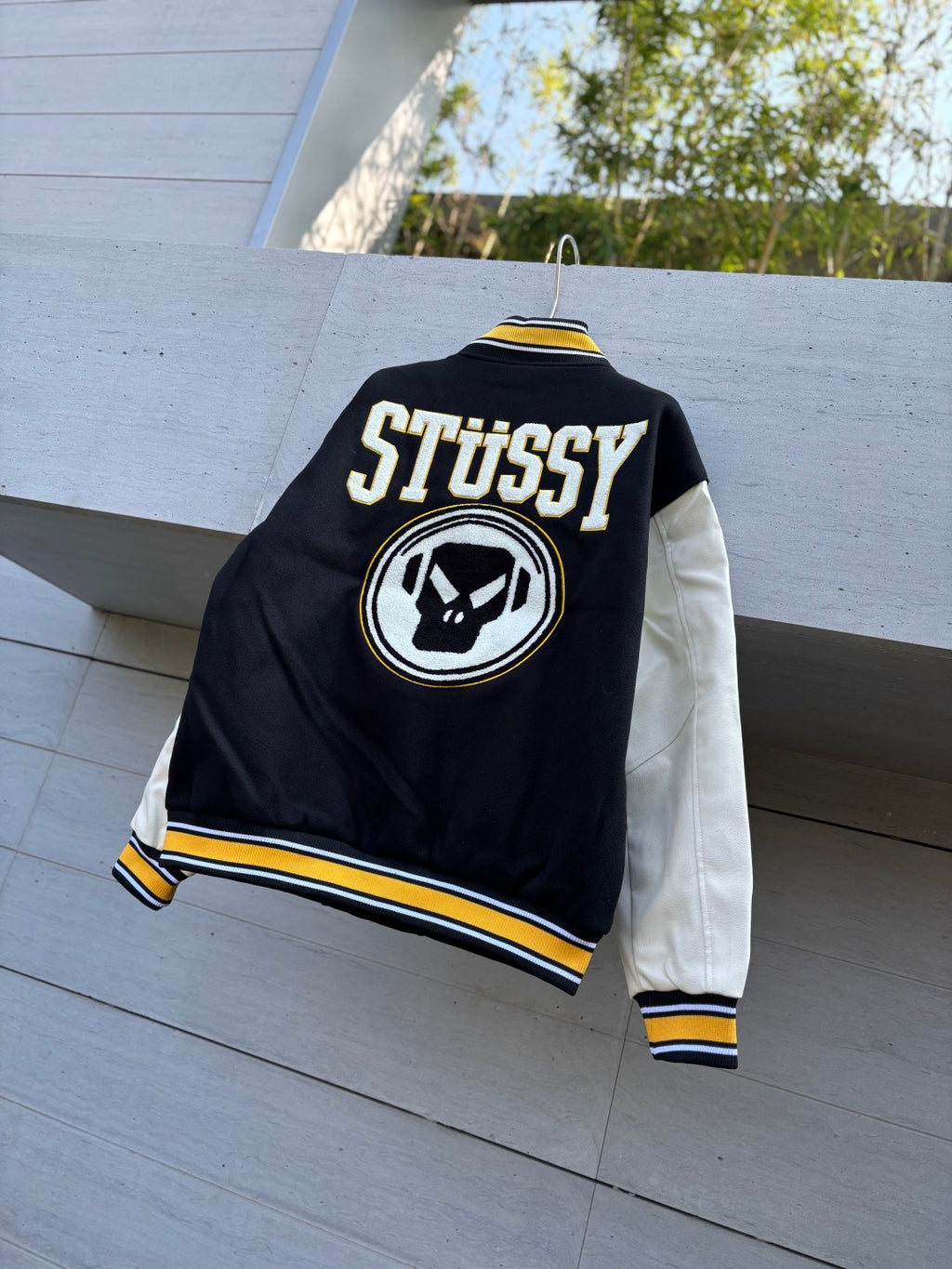 Black varsity jacket back view with STUSSY text, circular skull logo patch and yellow trim