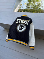 Black varsity jacket back view with STUSSY text, circular skull logo patch and yellow trim