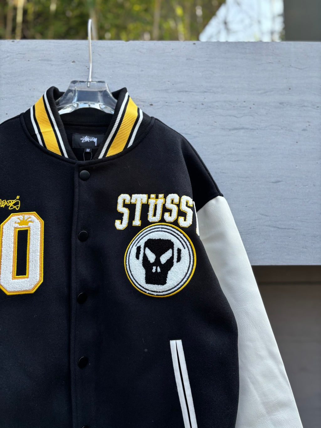 Black varsity jacket front close-up with STUSSY text, circular skull logo patch
