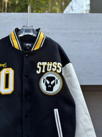 Black varsity jacket front close-up with STUSSY text, circular skull logo patch