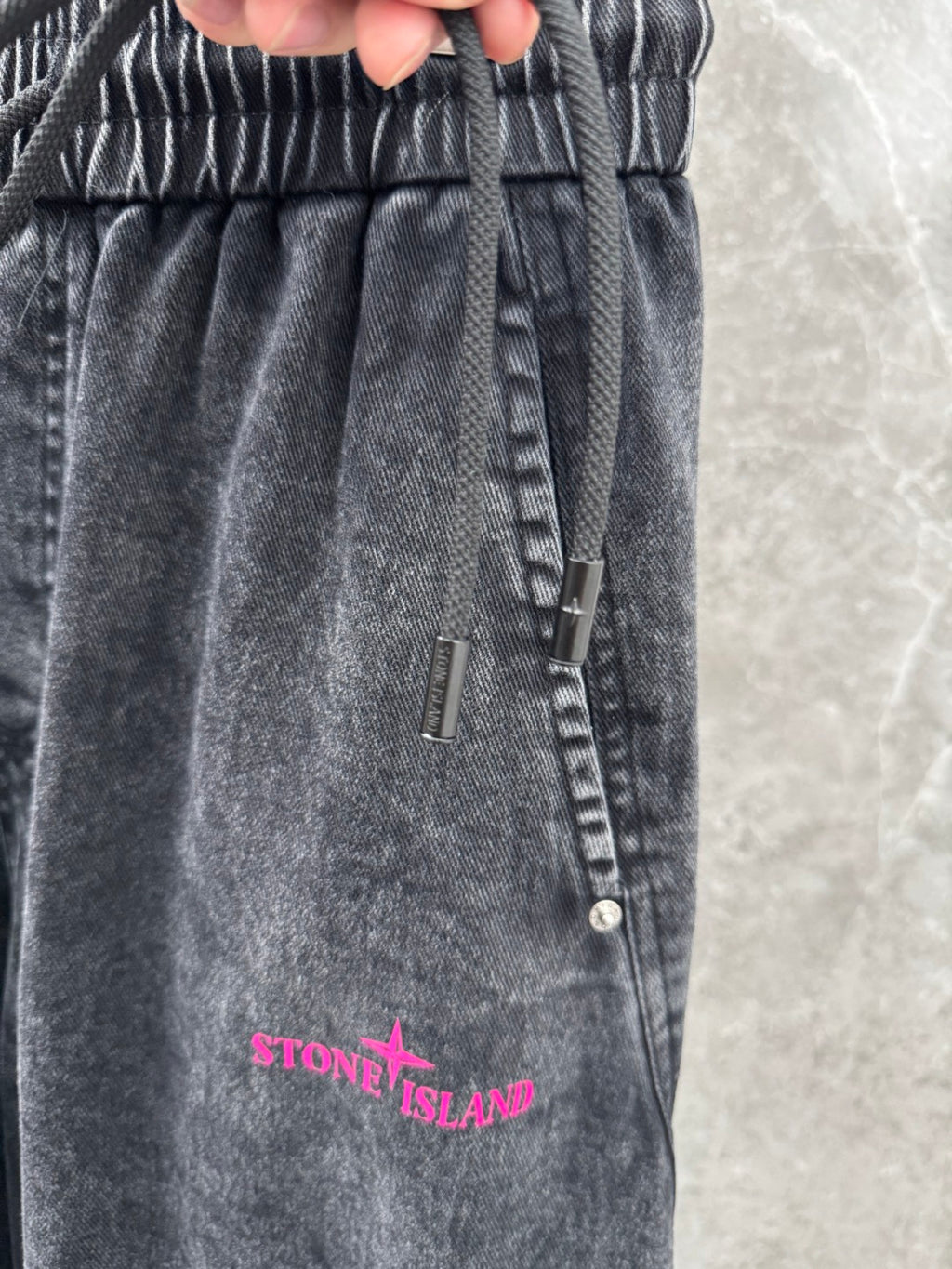Washed gray denim pants close-up: elastic waistband, drawstring, and pink Stone Island text.
