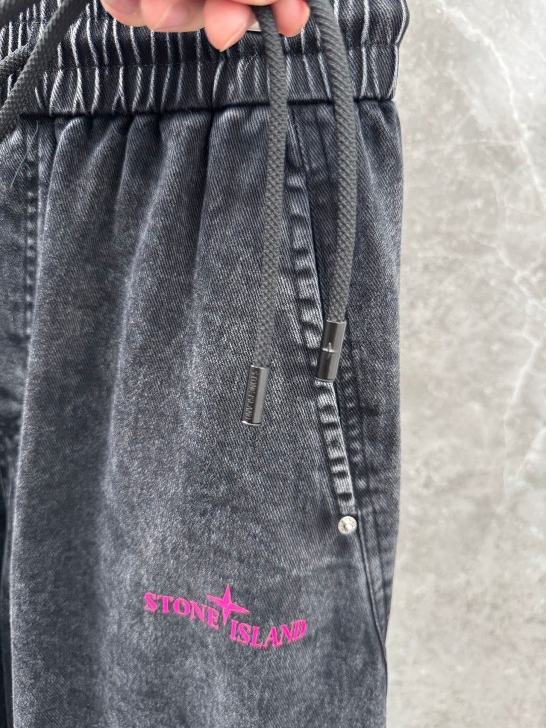 Washed gray denim pants close-up: elastic waistband, drawstring, and pink Stone Island text.