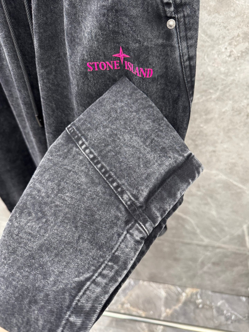 Washed gray denim pants detail with pink “STONE ISLAND” logo on front pocket area.