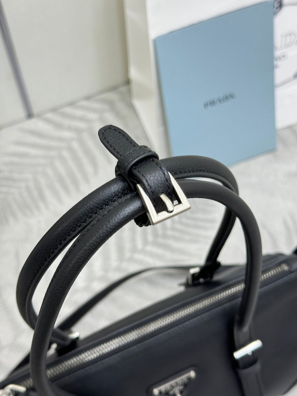 Prada handbag detail: dark leather strap and gold-tone buckle, textured top zipper.