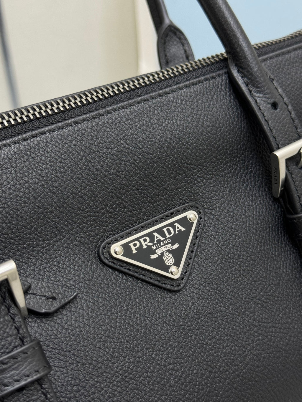 Prada handbag close-up: textured dark leather and metal logo plaque on front.