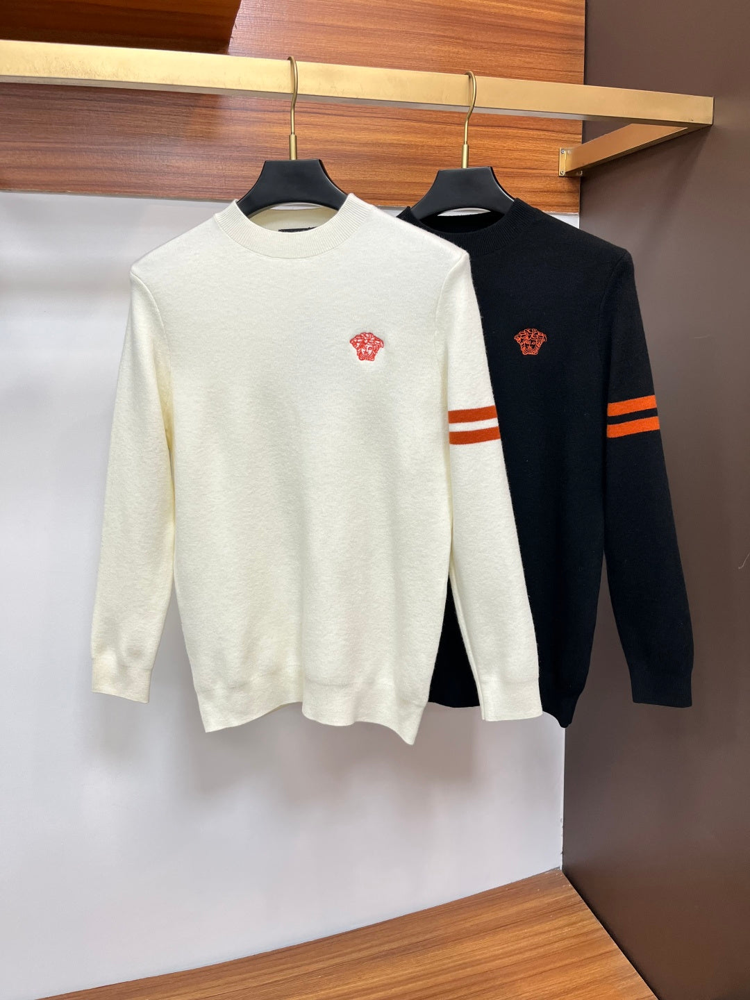 Cream and navy Versace crewneck sweaters side by side with red logo and sleeve stripes.