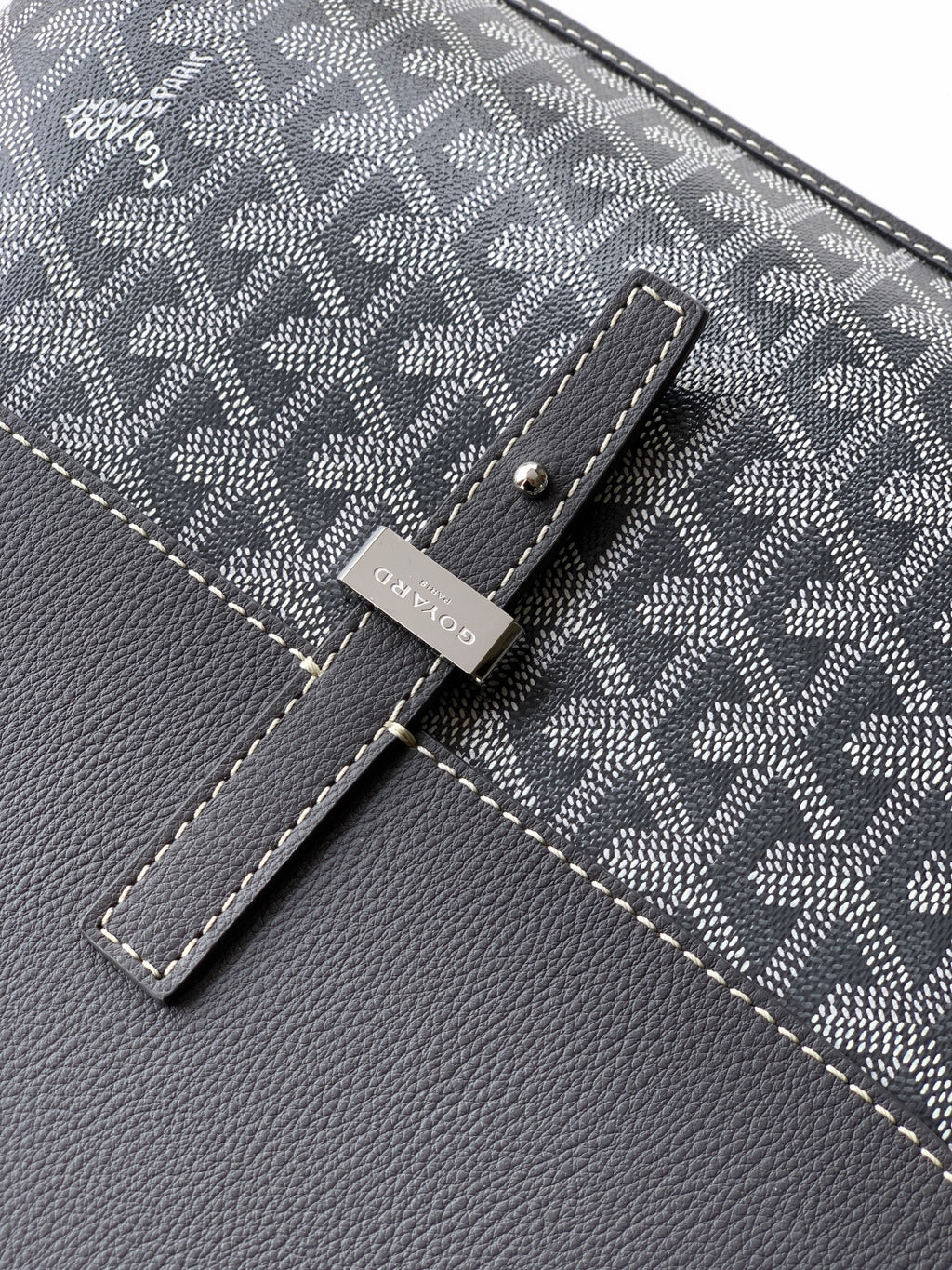 Goyard bag logo tag detail on gray patterned fabric with stitched leather strap tab