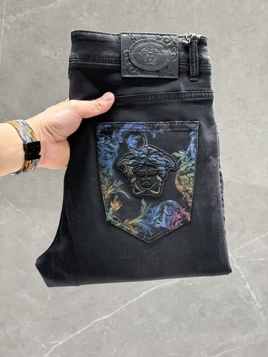 Dark denim jeans with a leather-look belt, button fly, and blue graphic back pocket patch.