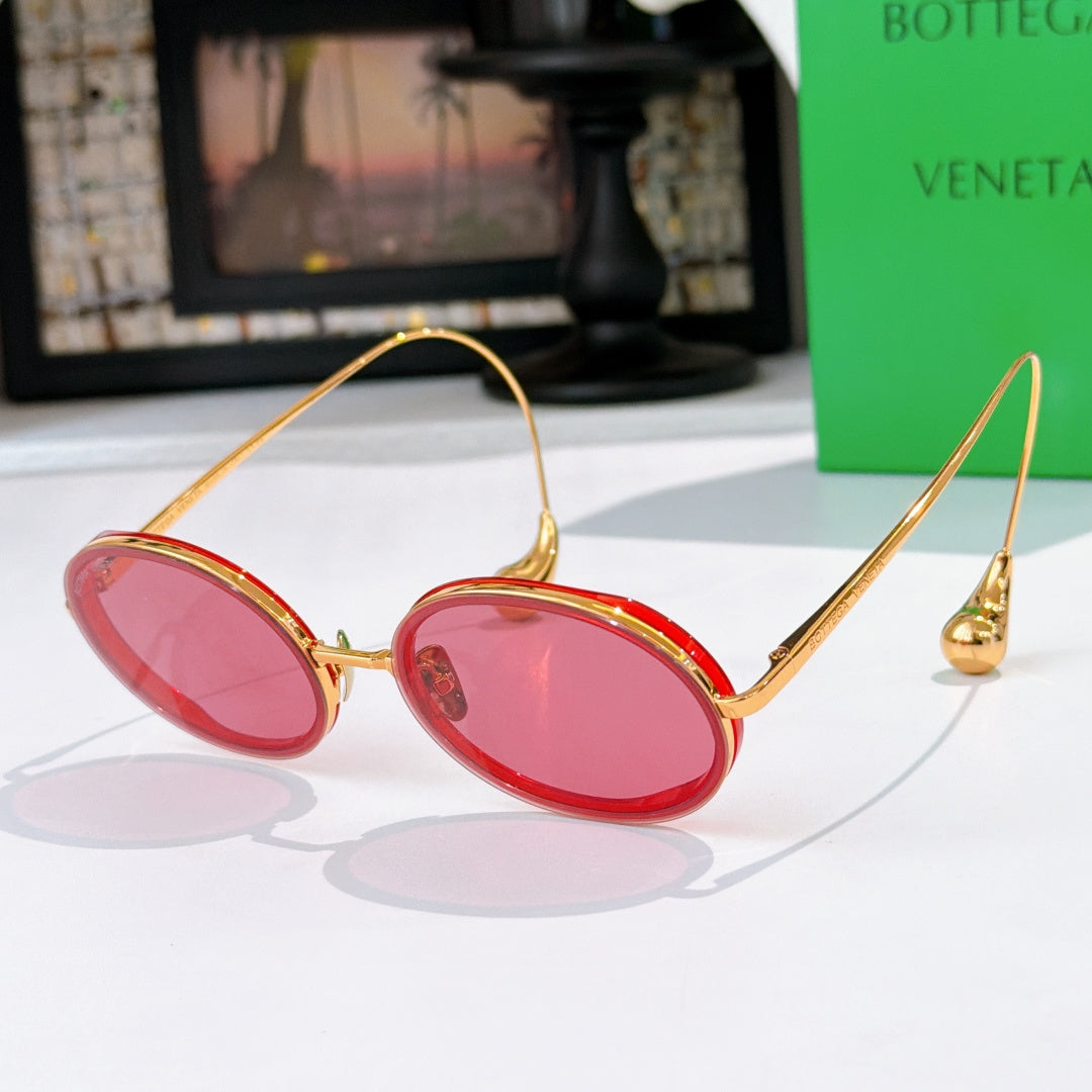 Oval sunglasses with pink tinted lenses and red frames with gold-tone metal accents.