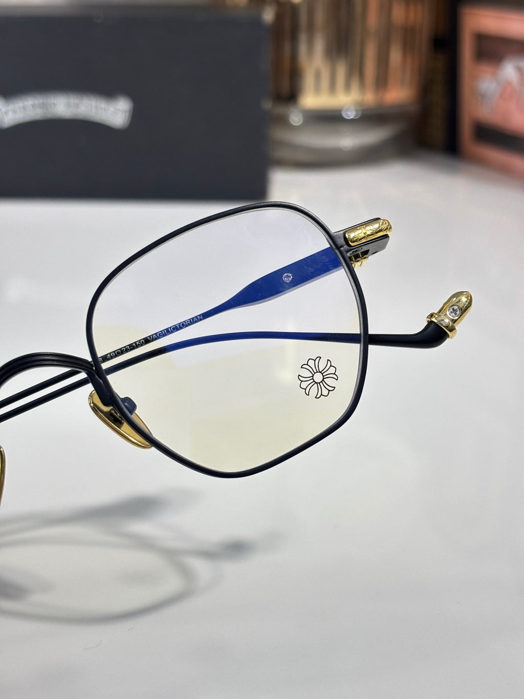 Black and gold round eyeglasses with blue stripe lens detail and flower emblem.