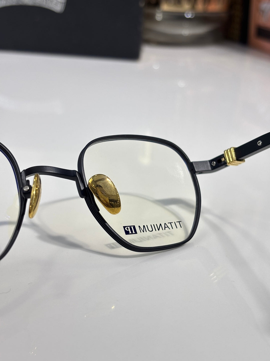 Close-up of black round eyeglass frame with gold temple and inner lens mark.
