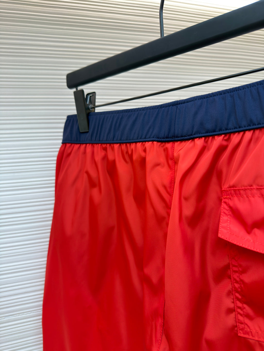 Red swim shorts with navy waistband and side pocket flap, close-up on hanger