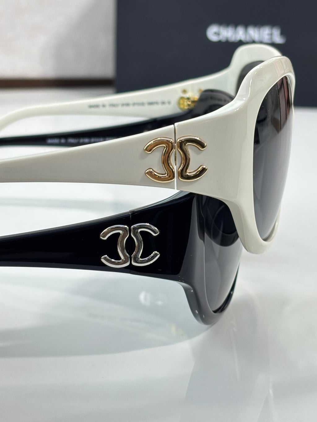 White sunglasses close-up with gold CC emblem on temple and dark lens