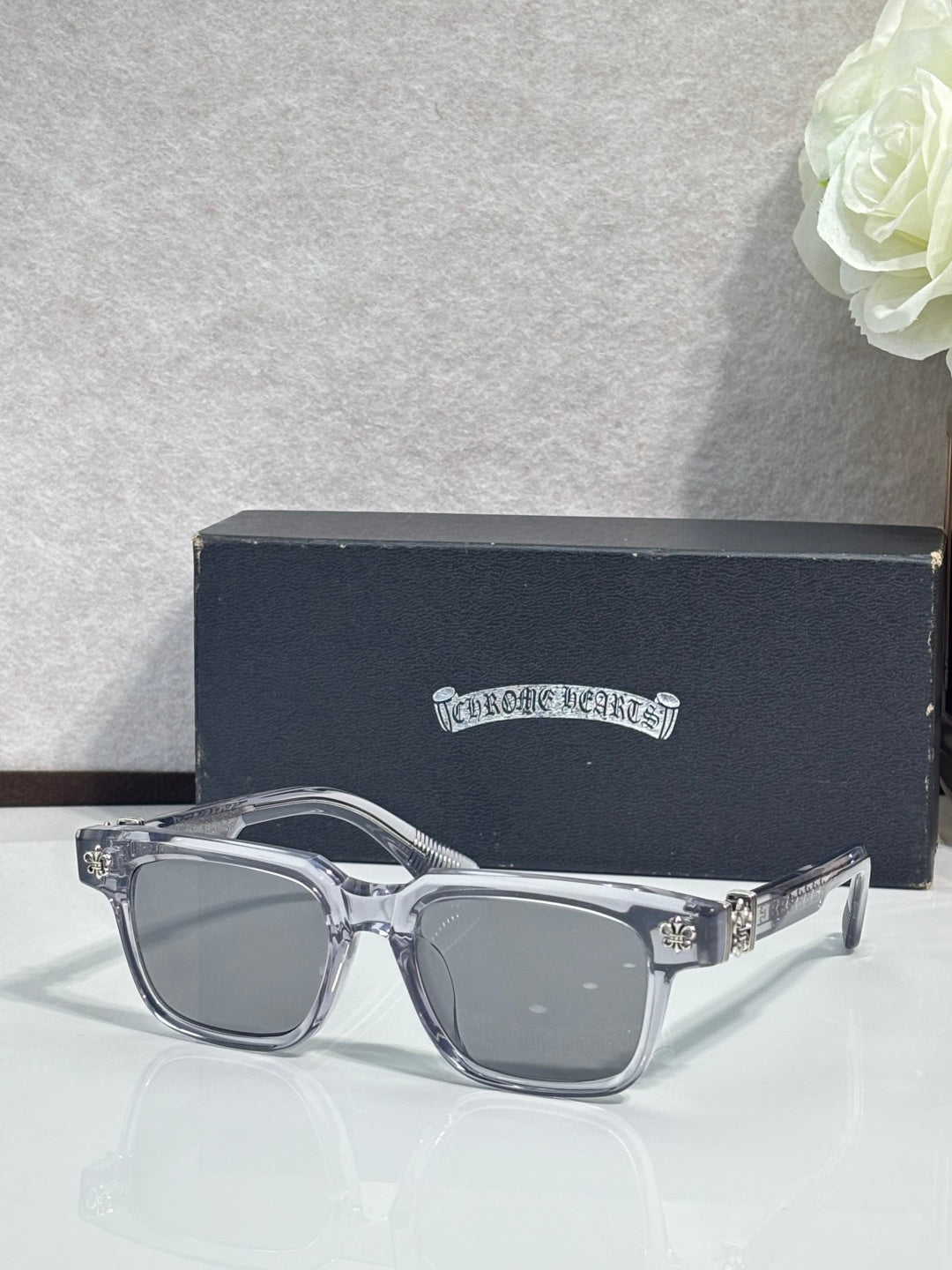 Clear-framed sunglasses with chrome finish and small gold fleur-de-lis details