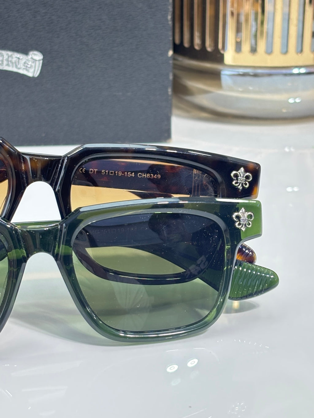 Green and brown sunglasses with chrome frame, dark lens, and fleur-de-lis detail