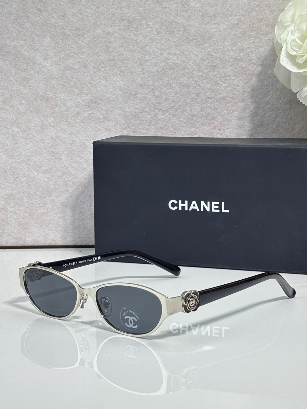 Oval sunglasses with silver frame, dark lenses, and gold bow medallion