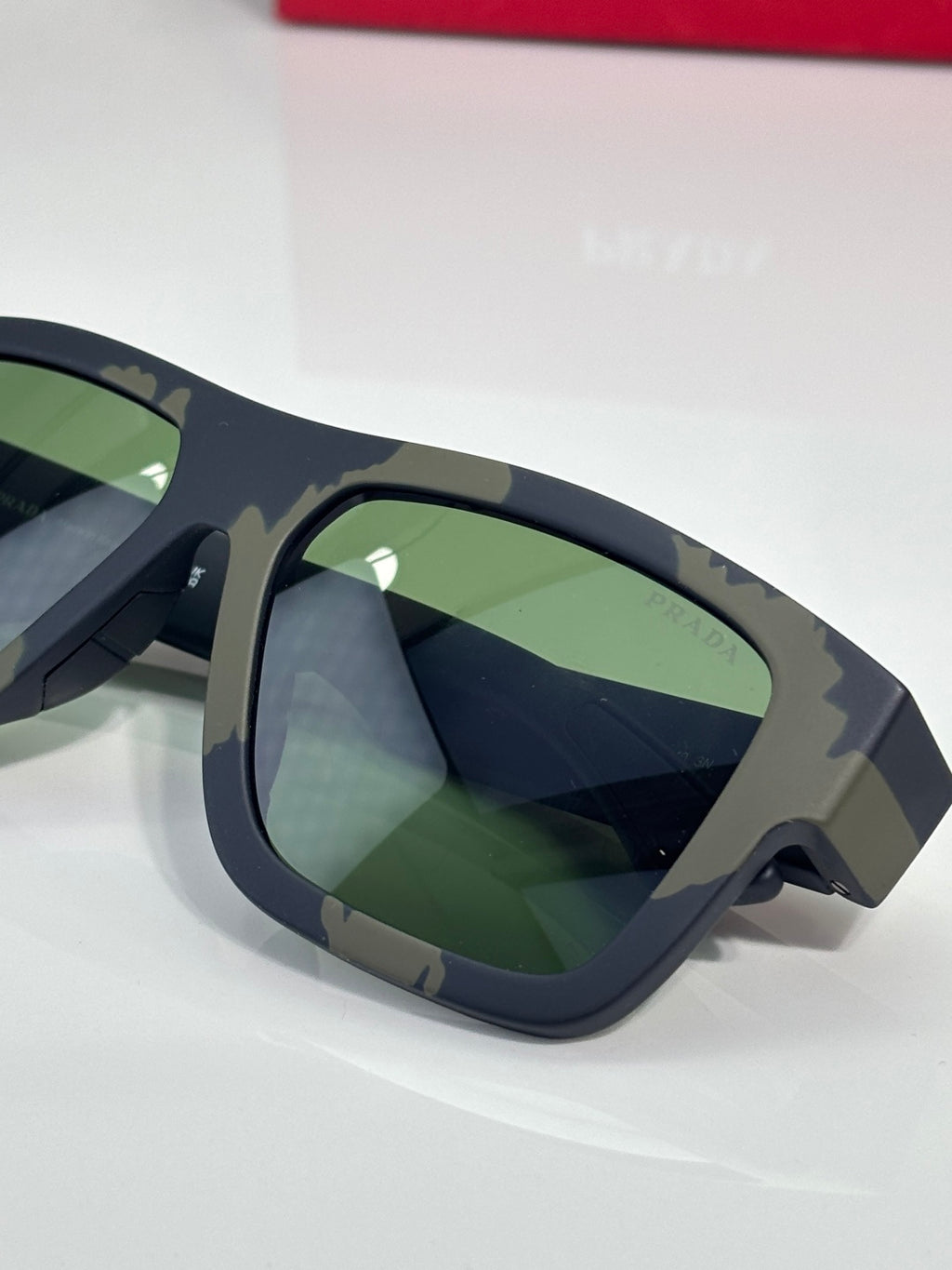 Camo sunglasses side close-up with green lenses and Prada red logo on arm.