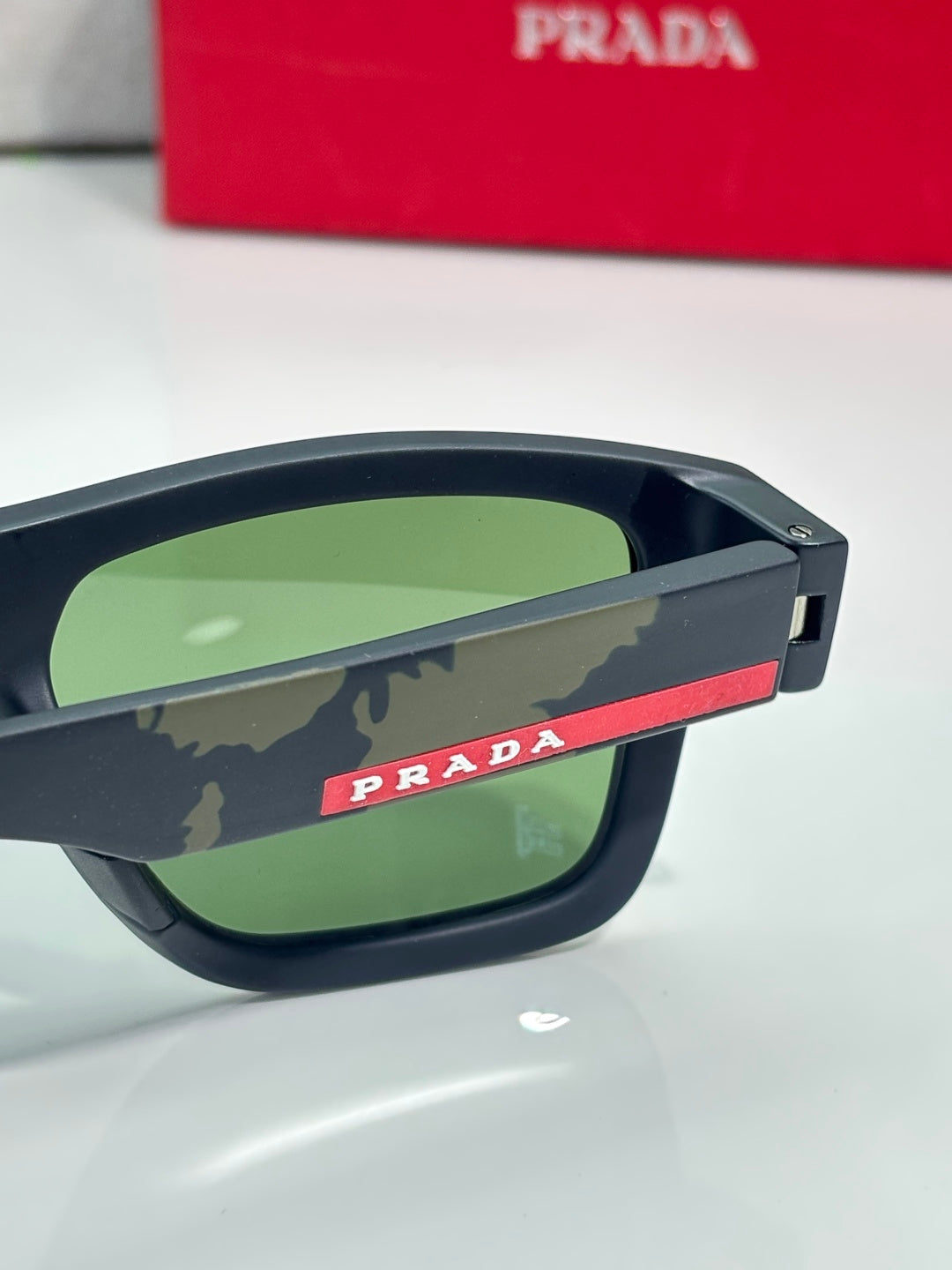 Camo sunglasses close-up: green gradient lenses, temple with Prada red stripe.