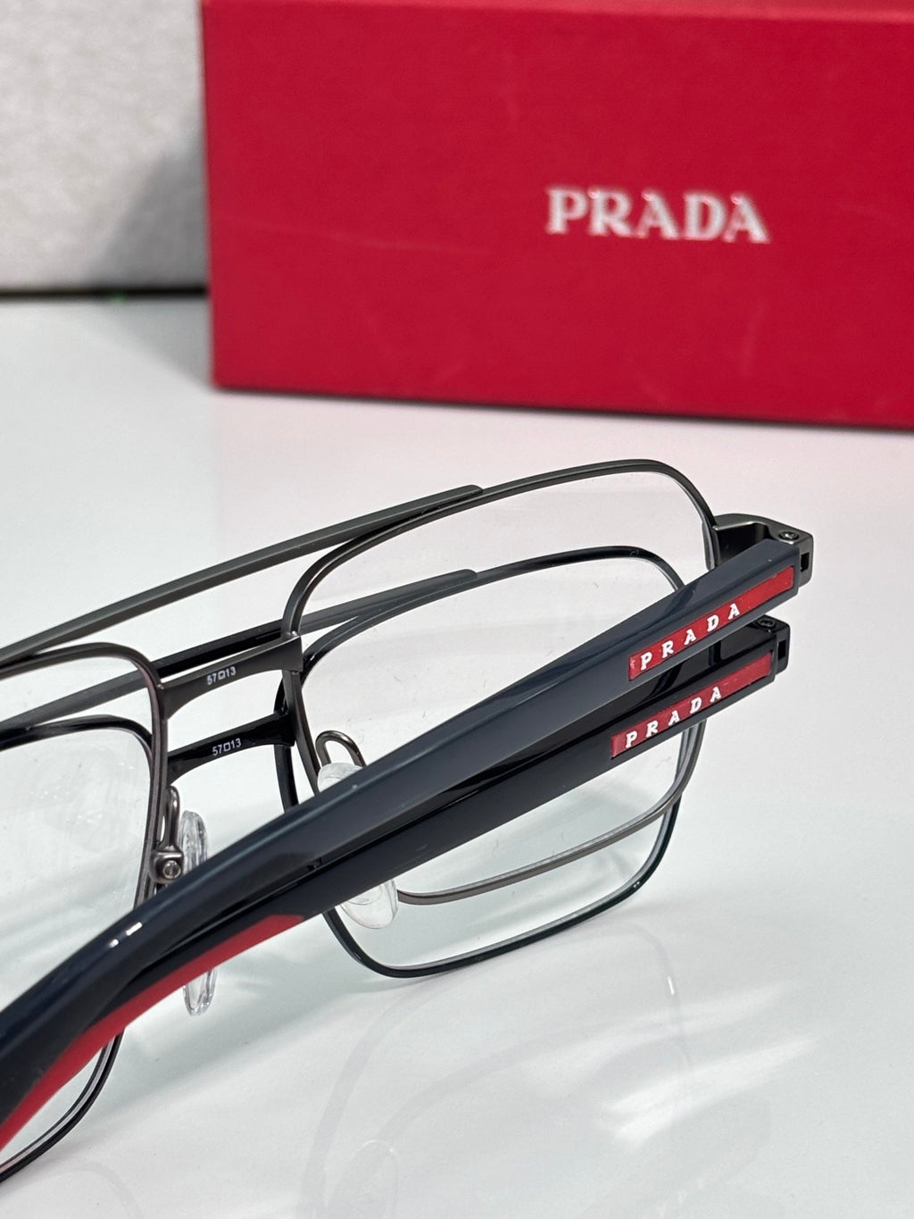 Prada eyeglasses close-up: black temples with red “PRADA” text and clear lens front.