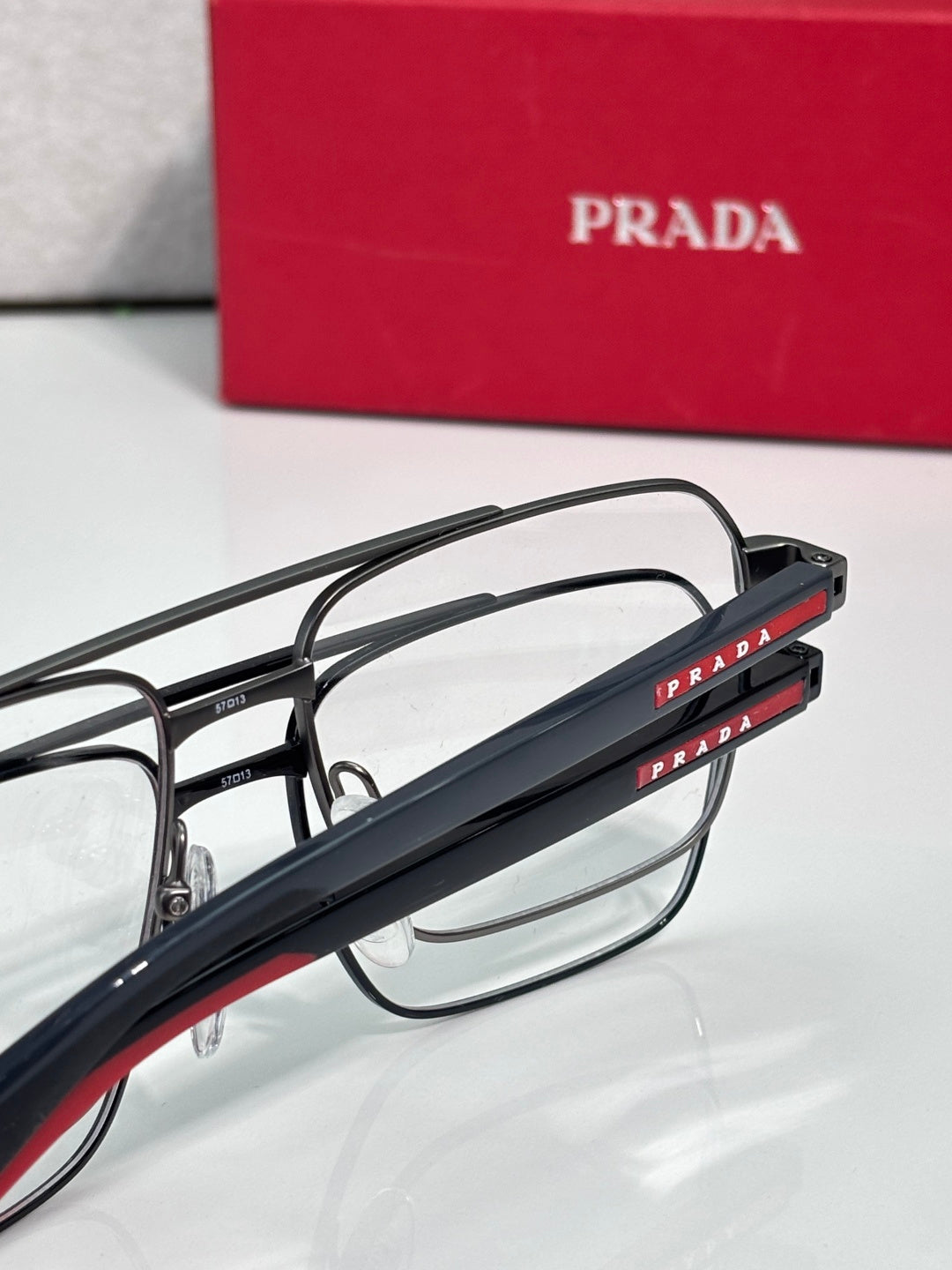 Prada eyeglasses close-up: black temples with red “PRADA” text and clear lens front.