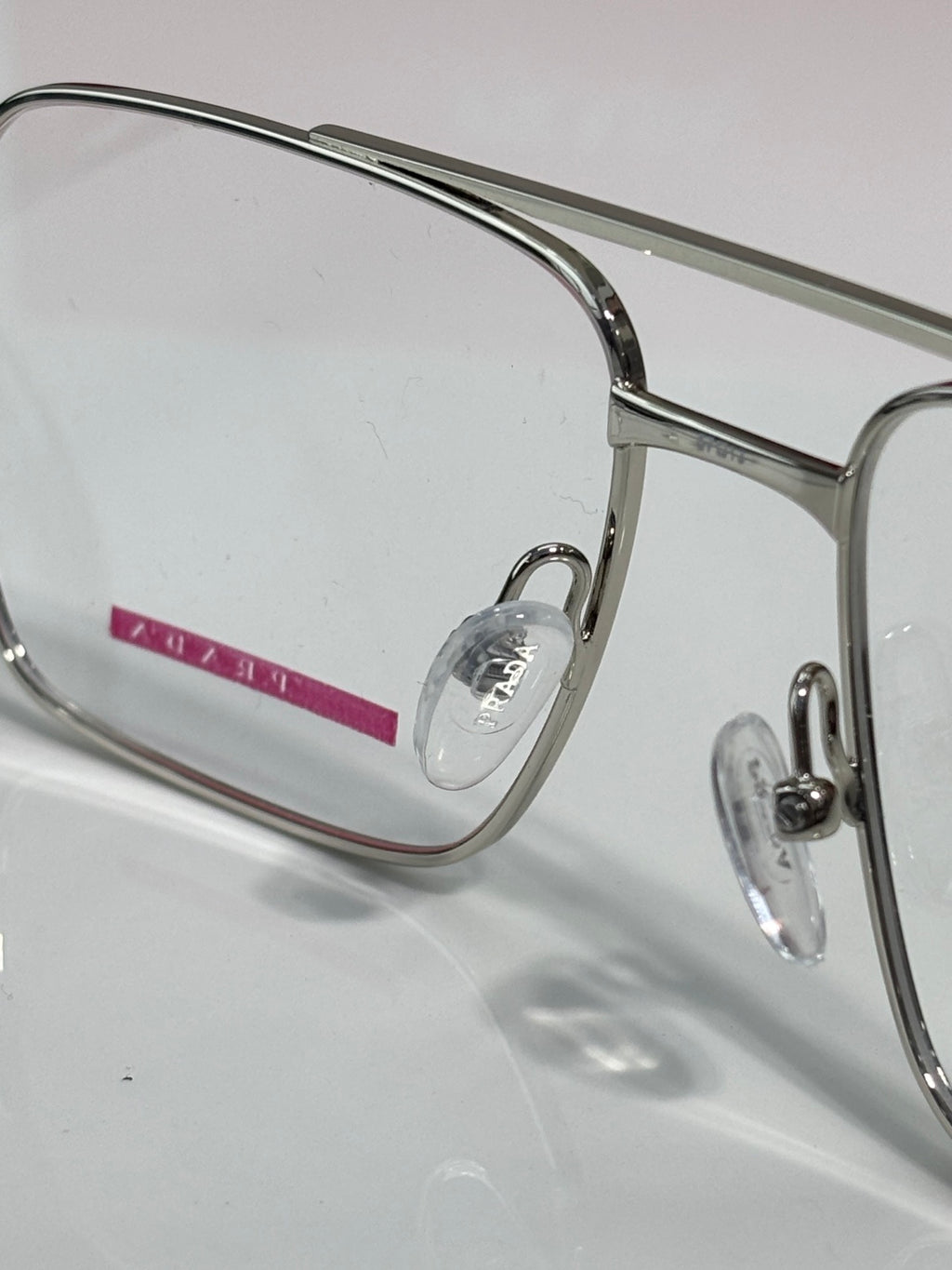 Prada eyeglasses close-up: clear lens and metal frame with small red logo detail.